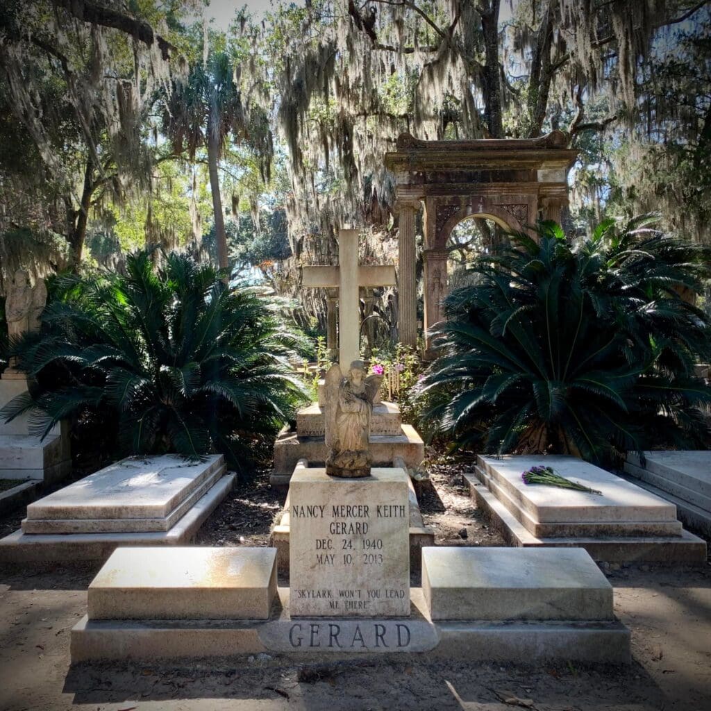 A grave marked for Nancy Mercer Keith Gerard, featuring a cross and an angel statue, is surrounded by lush greenery in the historic district of Savannah, Georgia.