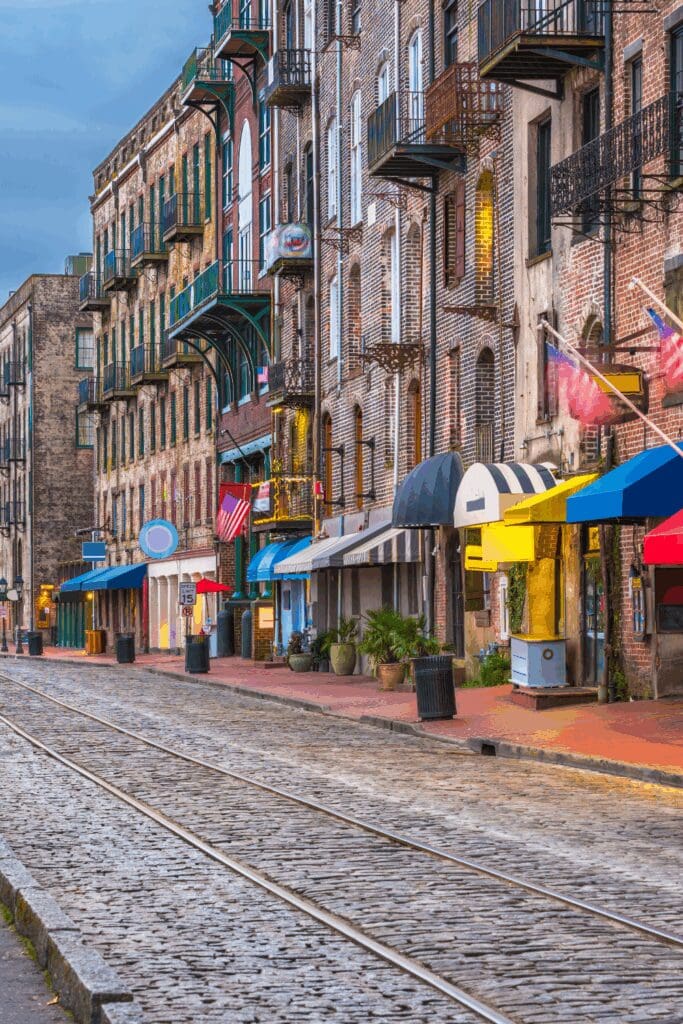 A cobblestone street lined with historic brick buildings featuring storefronts and balcony railings in the historic district of Savannah, Georgia.