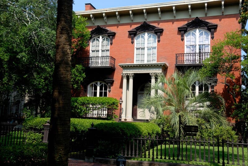 A historic brick building with decorative balconies and a columned entrance is surrounded by greenery and iron fencing in the historic district of Savannah, Georgia.